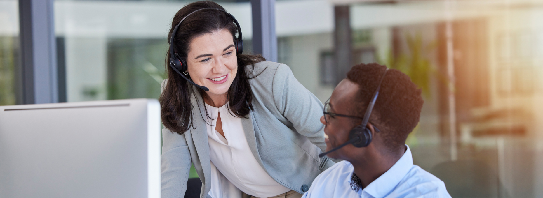 Smiling customer support agent in a headset leaning over to help a colleague at his computer in a bright office