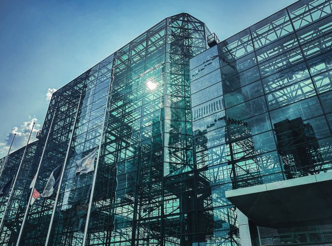 Glass facade of a modern building reflecting the sky, sun, and nearby flags. The structure has a geometric design, creating a sleek, futuristic vibe.