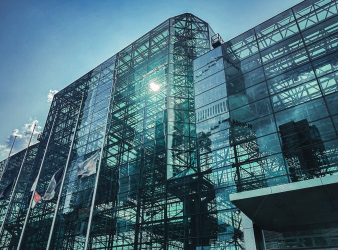 Glass facade of a modern building reflecting the sky, sun, and nearby flags. The structure has a geometric design, creating a sleek, futuristic vibe.