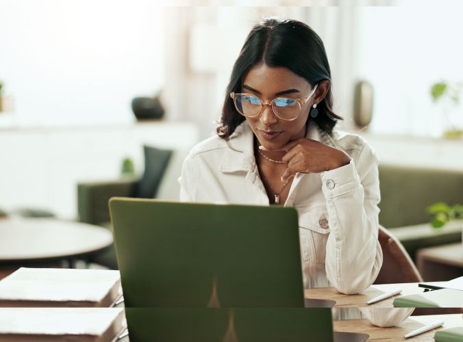 Woman in glasses and white jacket works on a laptop at a stylish home office. Sunlight softly illuminates the space, creating a calm and focused atmosphere.