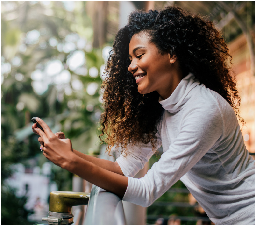 A smiling woman with curly hair leans on a railing, typing on her smartphone. She's outdoors, surrounded by blurred greenery, wearing a white top.
