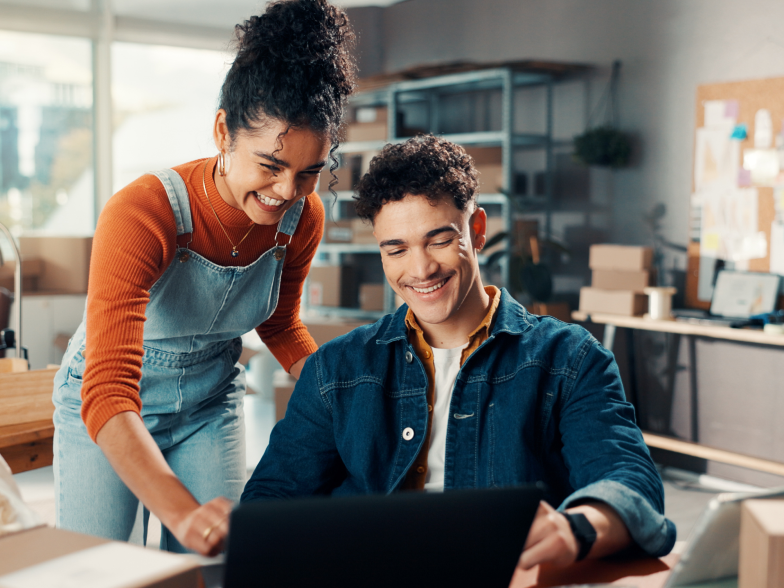 Two coworkers smiling while they review something on a laptop together in a small ecommerce stockroom with shelves and shipping boxes