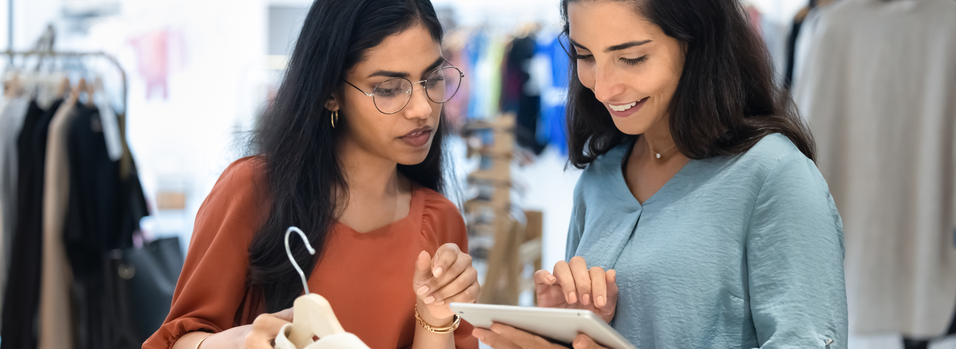 Retail associate checks a customer’s details on a tablet while helping a smiling shopper who’s holding a white blouse in a clothing store