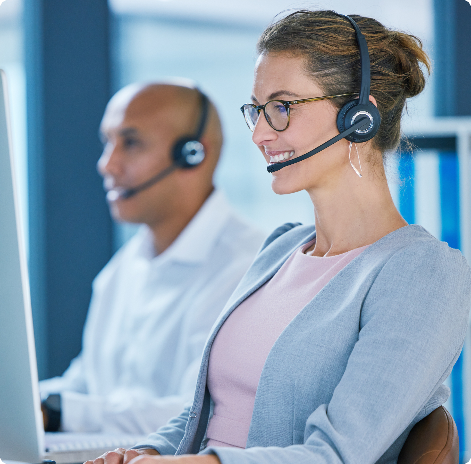 A woman in glasses and a headset smiles, working at a computer. In the background, a man with a headset sits at another desk. The scene is professional and focused.