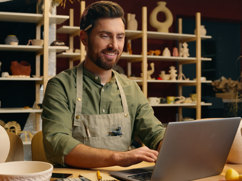 Smiling small-business owner in a canvas apron works on a laptop in his ceramics shop, with shelves of pottery and vases behind him