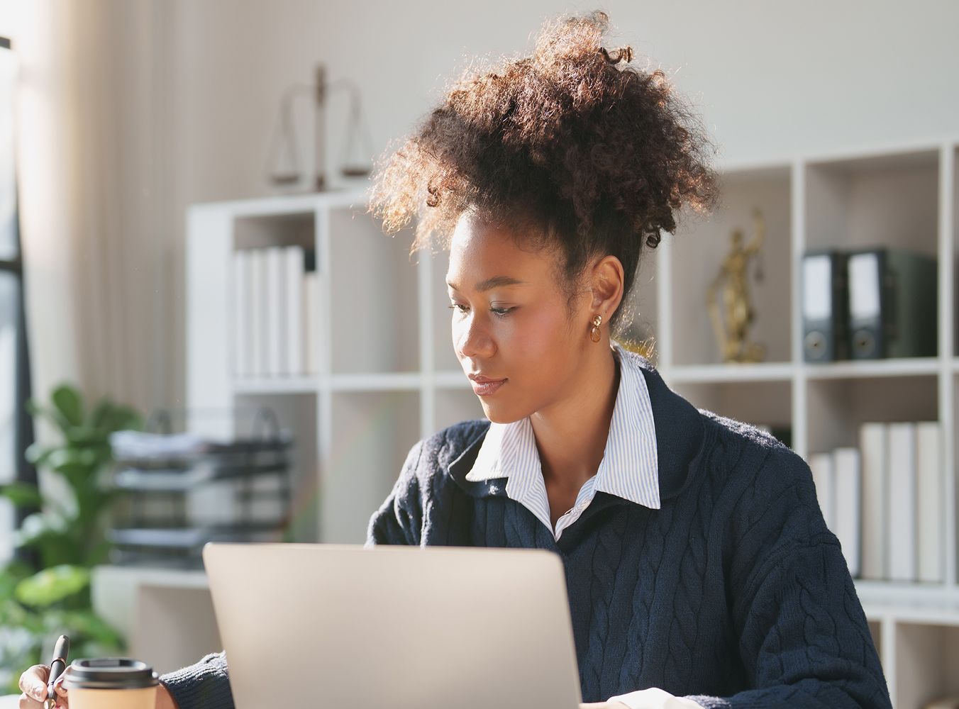 A focused woman in a navy sweater works at a laptop in a bright office, with bookshelves behind her and a coffee cup nearby, conveying concentration.