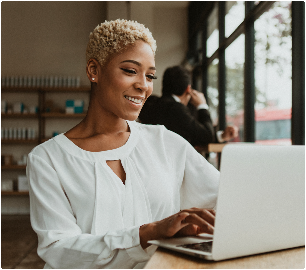 A woman with short curly hair smiles while using a laptop in a bright cafe. She wears a white blouse. A person is seated in the background by large windows.