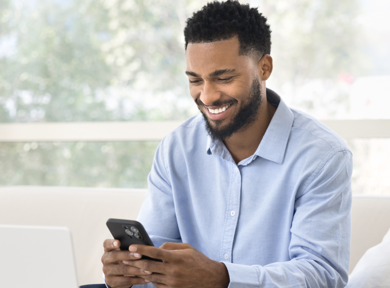 Smiling man in a light blue shirt uses a smartphone indoors, seated comfortably with a blurred nature background. He appears relaxed and happy.