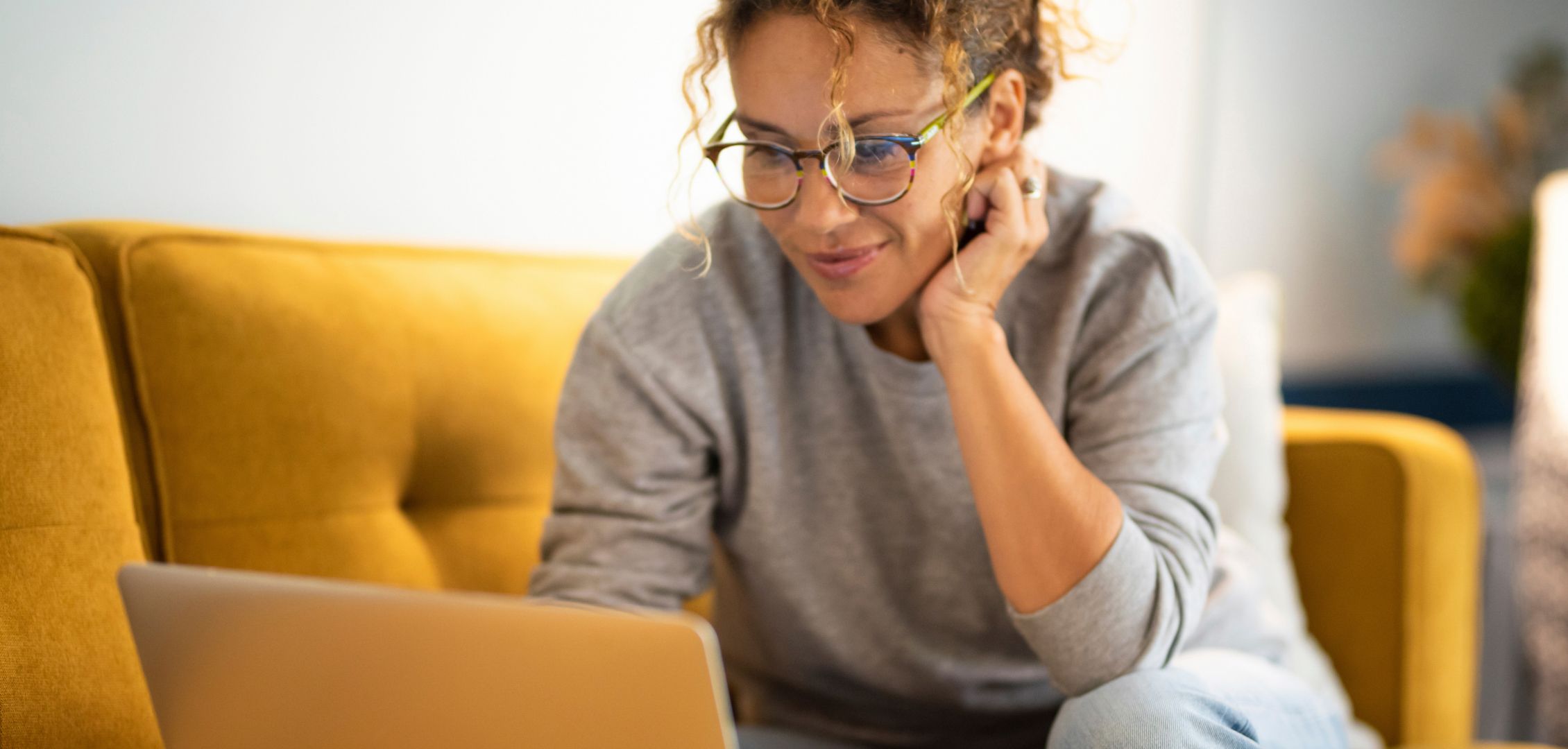 A person with curly hair and glasses smiles while using a laptop on a yellow couch. They appear focused and relaxed in a cozy, bright room.