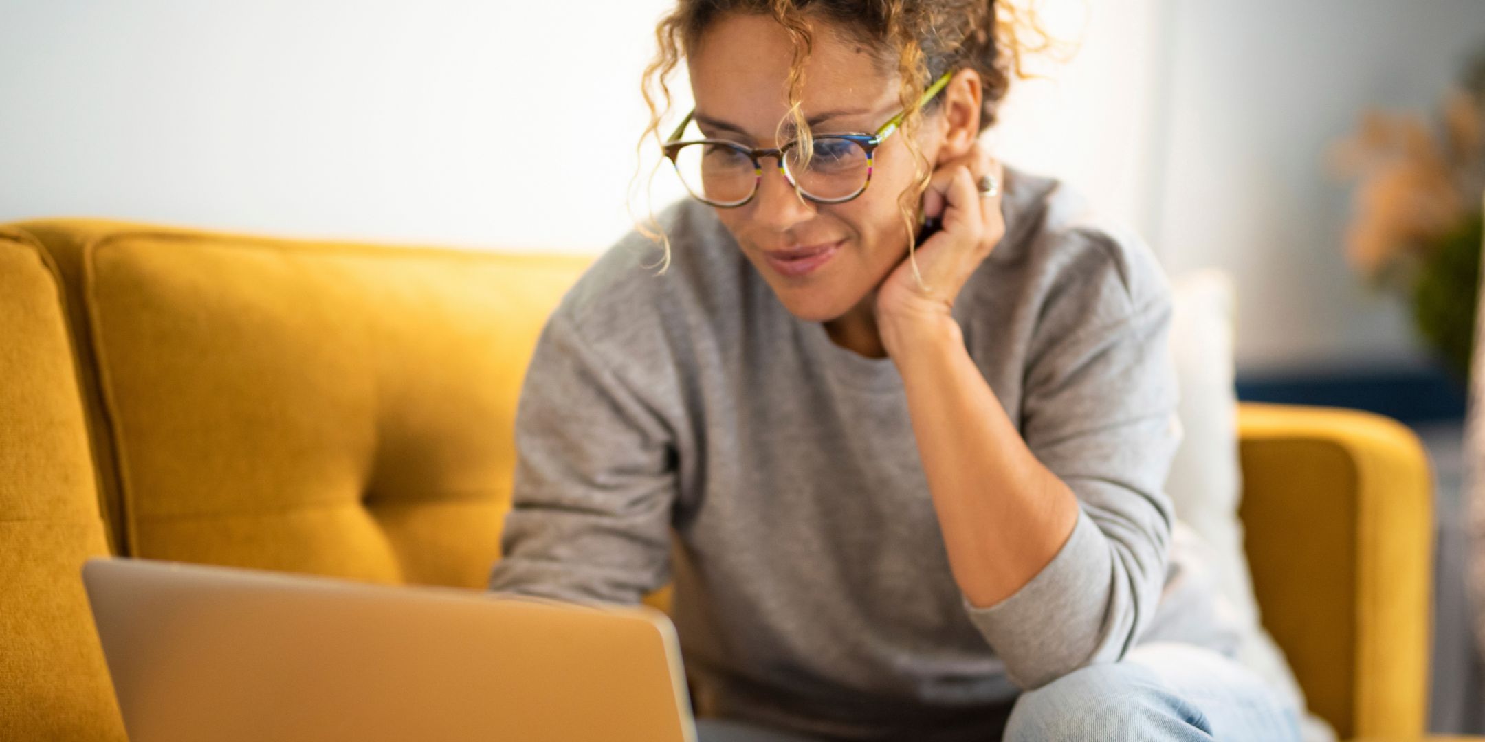 A person with curly hair and glasses smiles while using a laptop on a yellow couch. They appear focused and relaxed in a cozy, bright room.