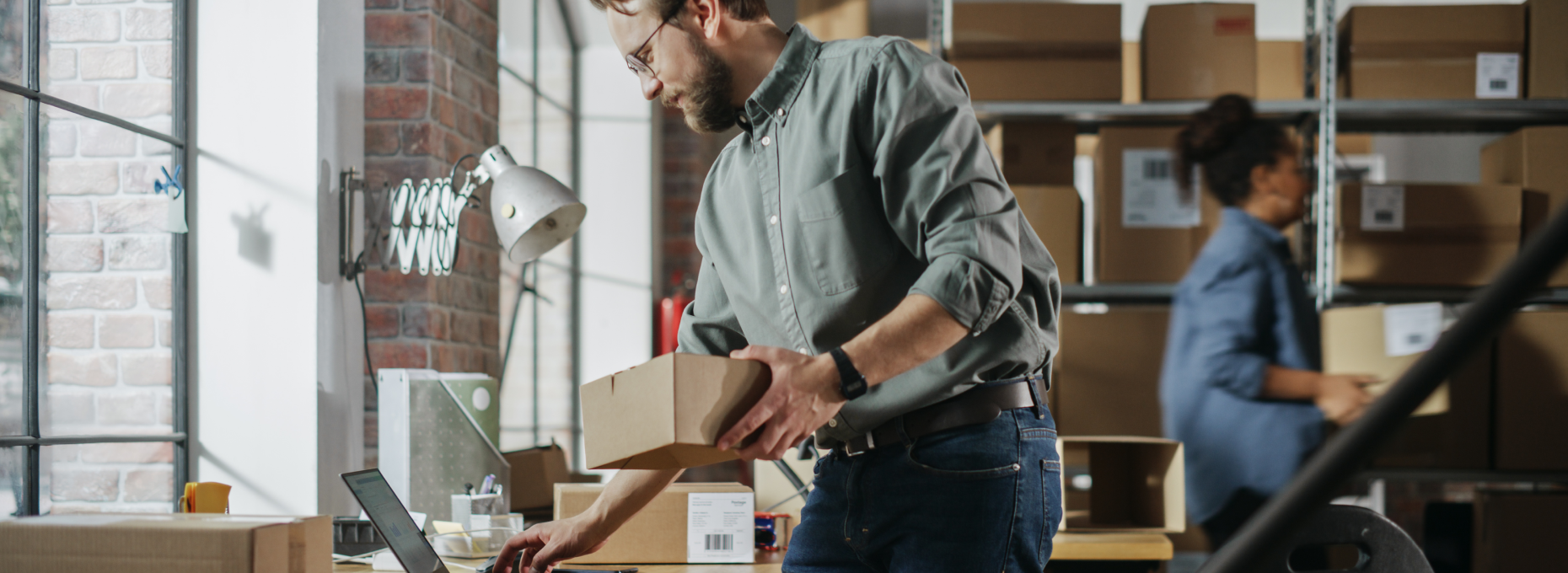 A small business owner checks his laptop while holding a package in a warehouse fulfillment area, with a colleague packing boxes in the background.