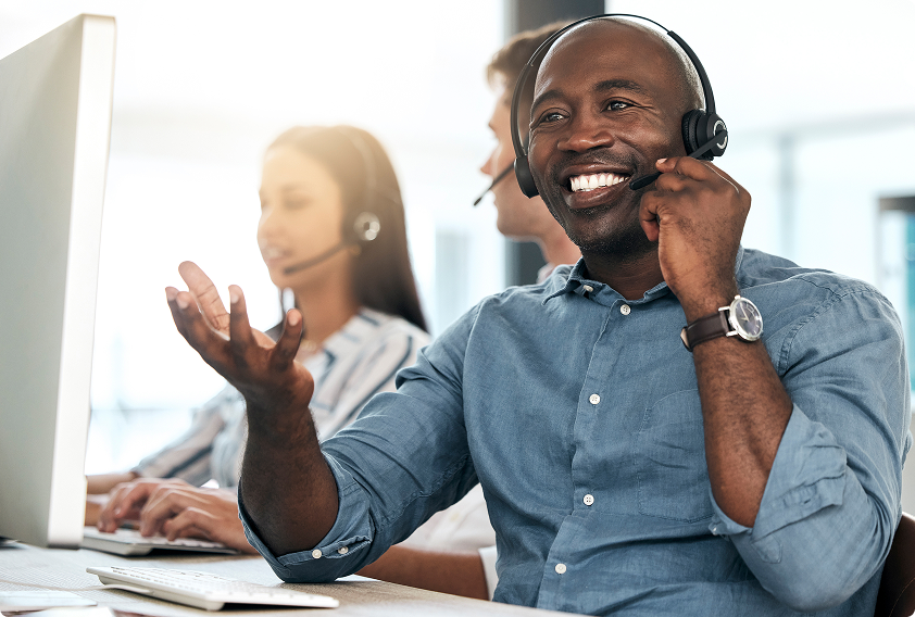 Smiling man in a blue shirt wearing a headset at a call center, gesturing while speaking. Two colleagues, also with headsets, work in the background.