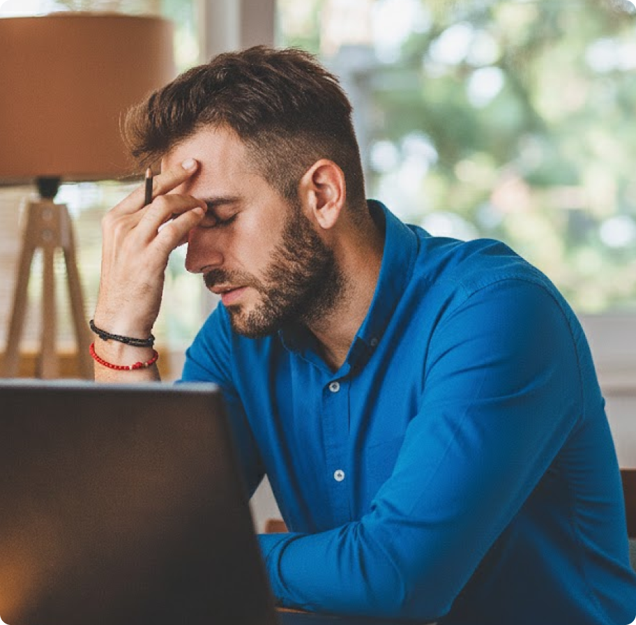 A man in a blue shirt sits at a desk with a laptop, looking stressed, holding his forehead. The room has soft lighting and a blurred view outside.