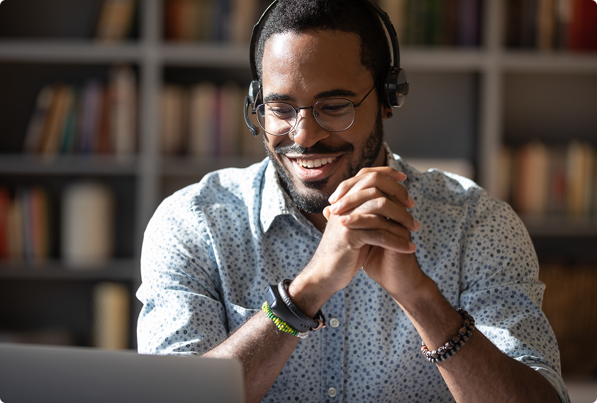 Smiling man with glasses and headphones, wearing a patterned shirt, sits with clasped hands in front of a laptop. Bookshelves in the blurred background.