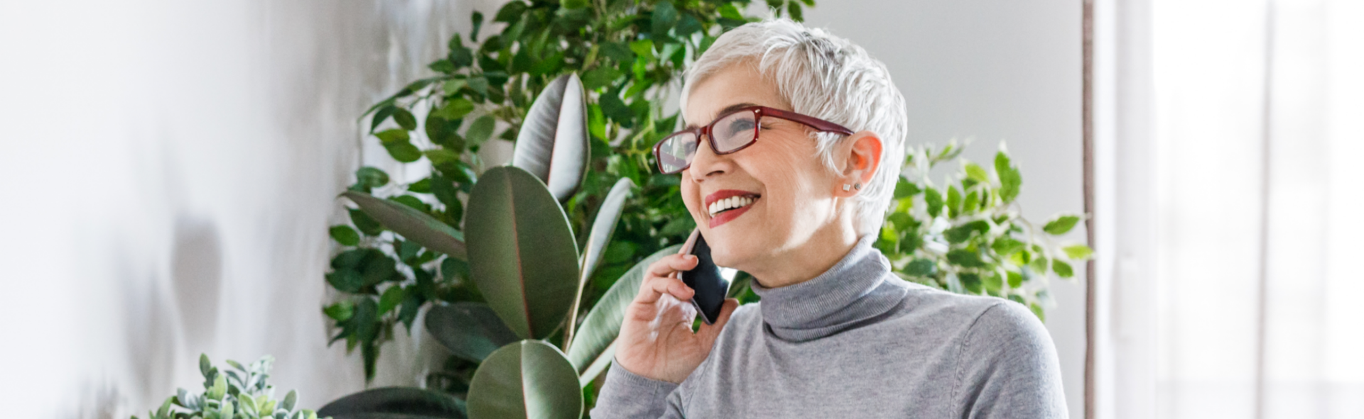 Older woman with glasses talking on a cell phone in front of indoor plants