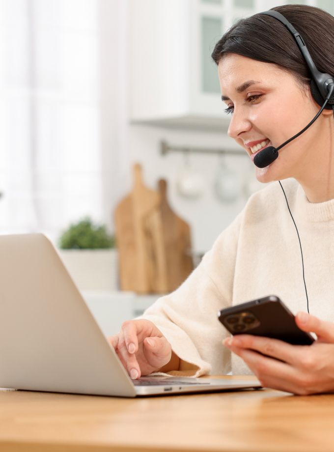 A woman in a headset smiles while working on a laptop, holding a smartphone in her other hand. The setting is a bright kitchen, conveying a cheerful, productive mood.