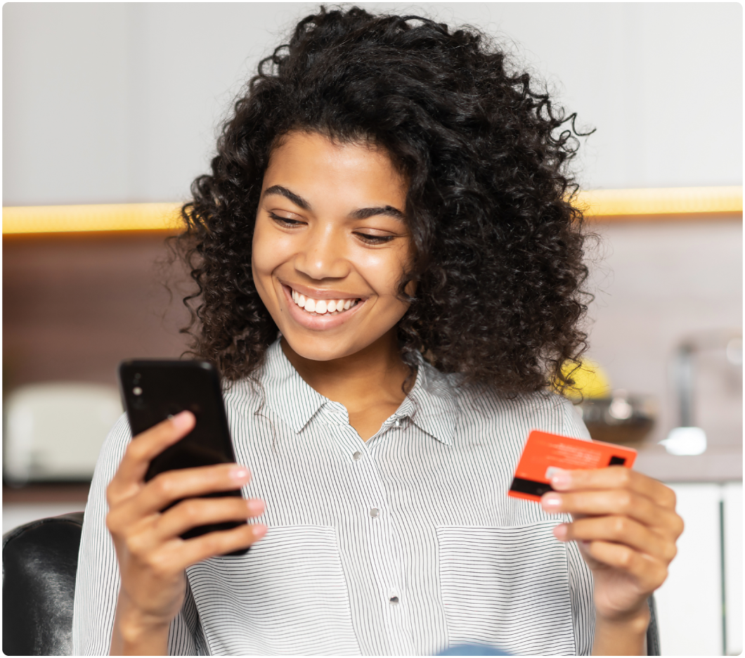 A smiling woman with curly hair holds a smartphone and a credit card, sitting in a bright room. She appears to be making an online purchase, conveying convenience.