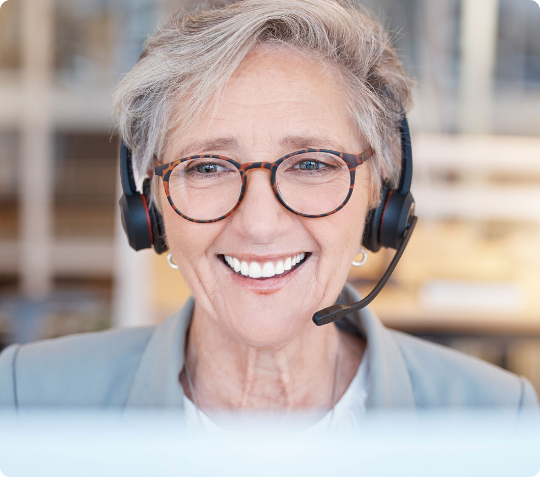 Smiling elderly woman wearing glasses and a headset, exuding warmth and professionalism in a bright office setting with blurred background.