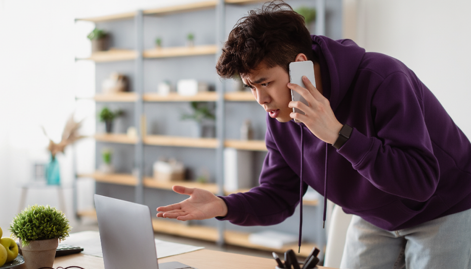 A man in a purple hoodie, looking frustrated, holds a smartphone to his ear while gesturing at a laptop. The setting is a modern home with shelves.