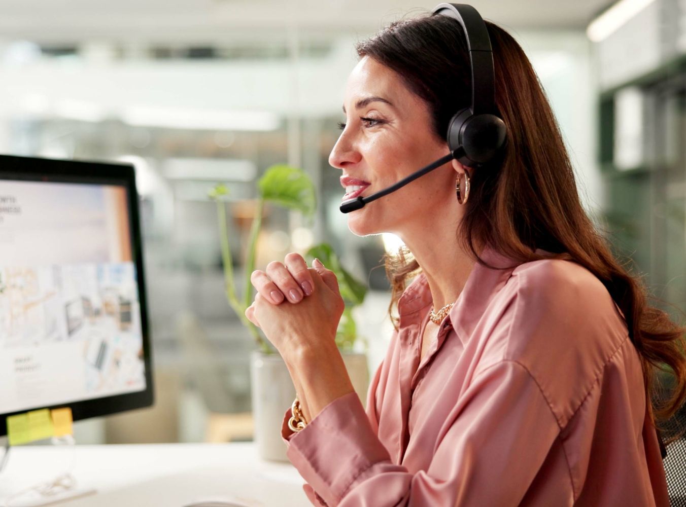 A woman in a pink blouse, wearing a headset, smiles while talking at a desk with a computer monitor. The office setting appears bright and modern.