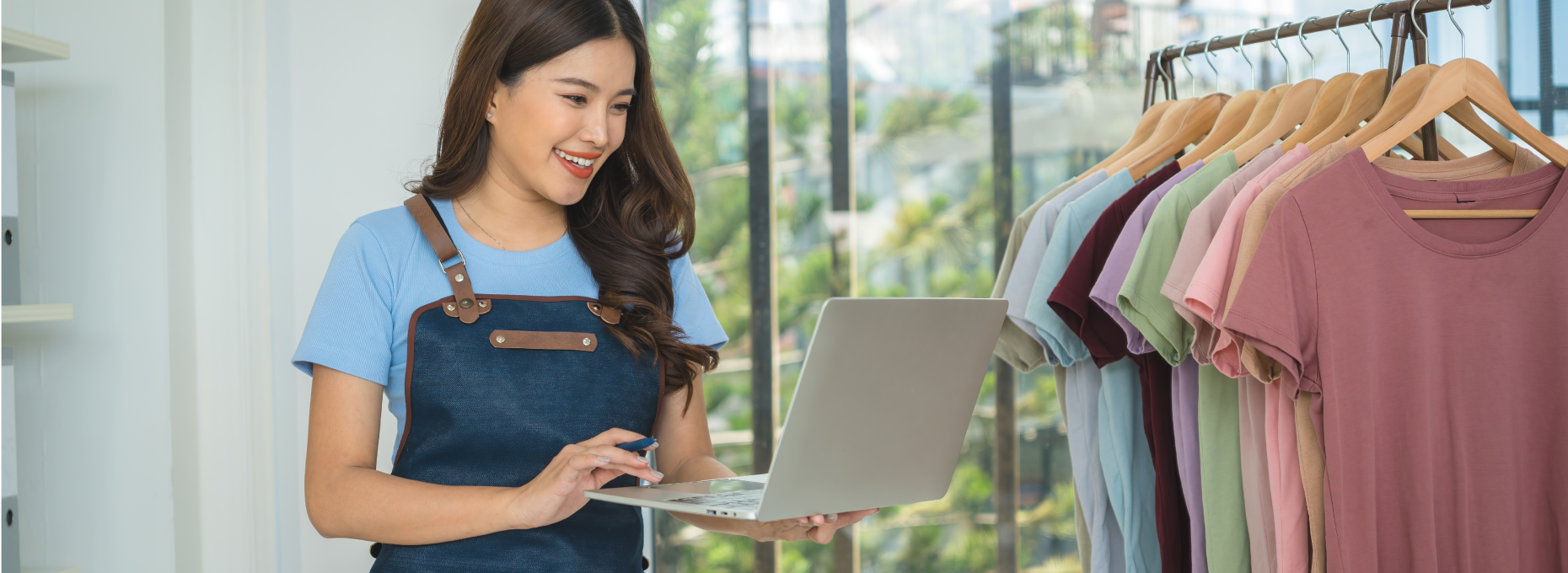 Boutique owner smiling at her laptop next to a rack of shirts and a packed shipping box