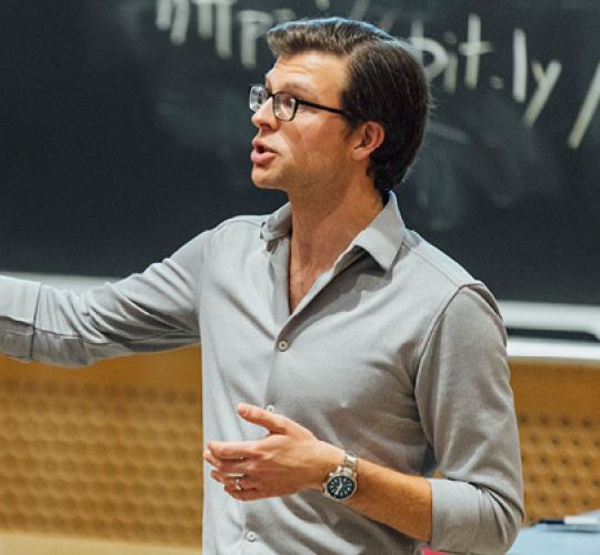 A man in glasses and a gray shirt is passionately lecturing in a classroom, gesturing with his hand. A chalkboard and link are visible behind him.
