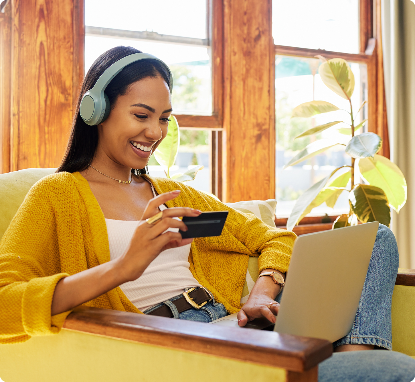 A woman in a yellow sweater and headphones smiles while online shopping with a credit card and laptop, sitting in a cozy chair by a sunlit window.