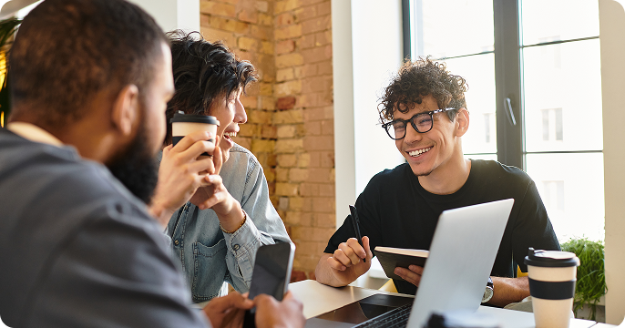 Three people sit around a table in a bright room with brick walls. They are smiling and chatting, holding coffee cups and notebooks, conveying a collaborative and friendly atmosphere.