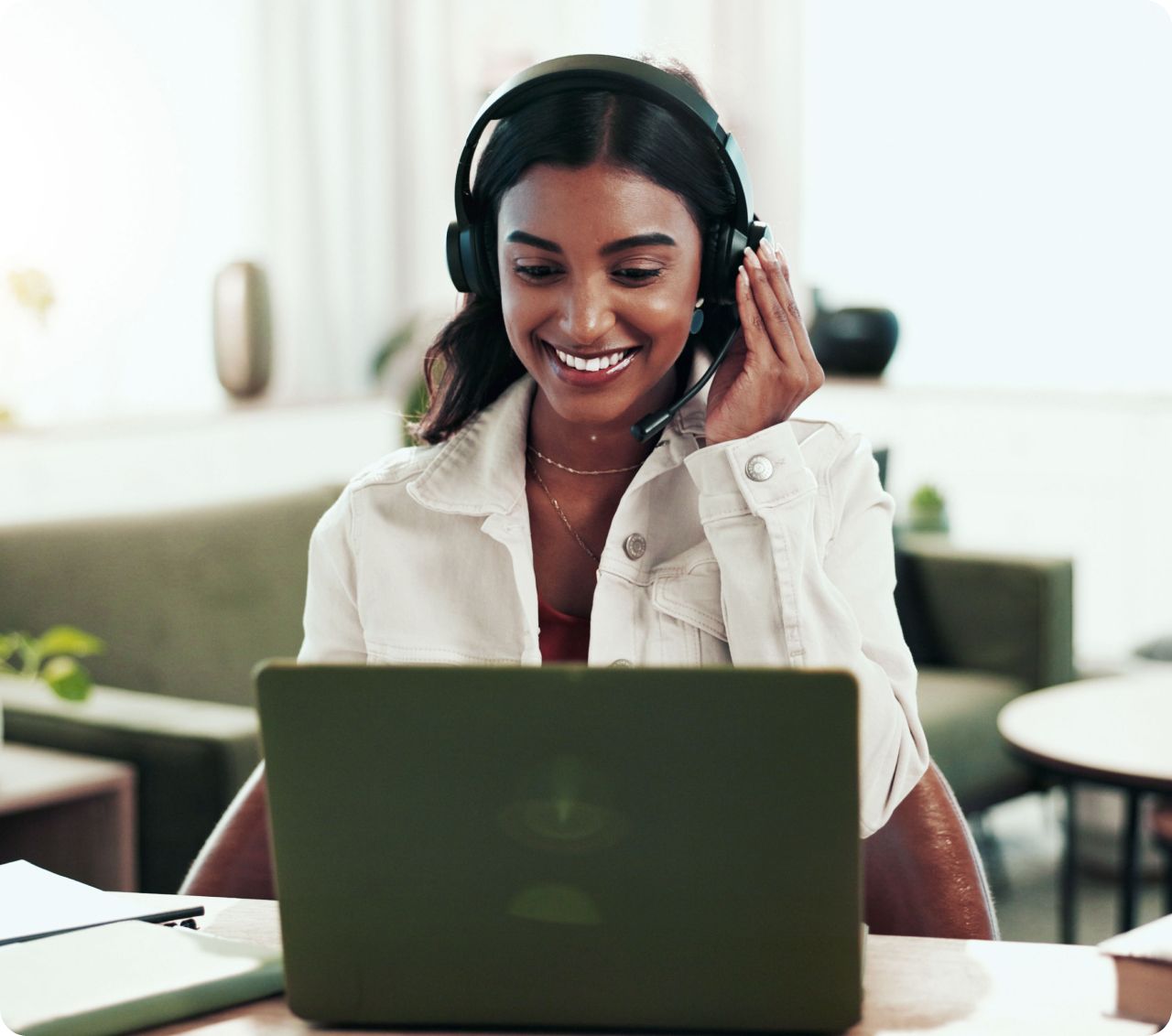 Smiling woman wearing a headset sits at a table with a laptop, engaging in a video call. The setting is a cozy room with soft lighting and neutral tones.