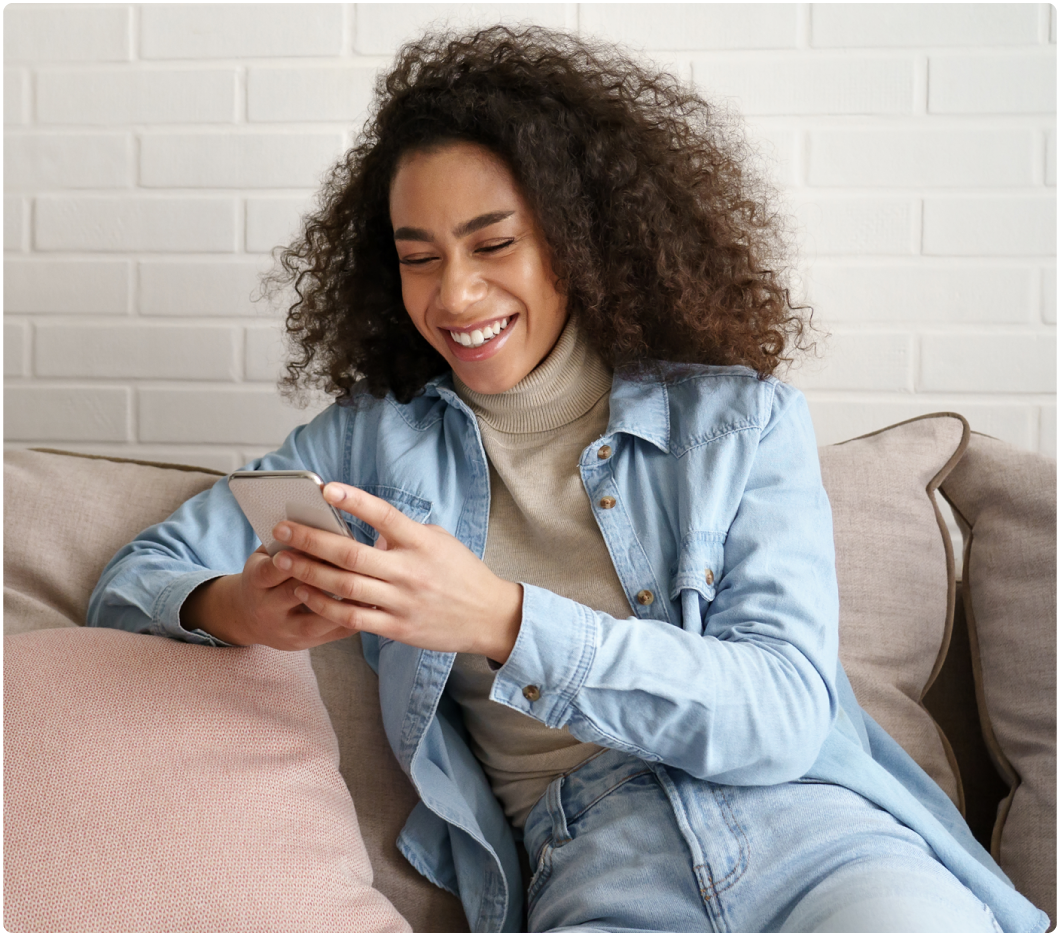 Smiling woman with curly hair sits on a beige sofa, looking at her phone. She wears a light blue jacket over a beige turtleneck, conveying a joyful mood.