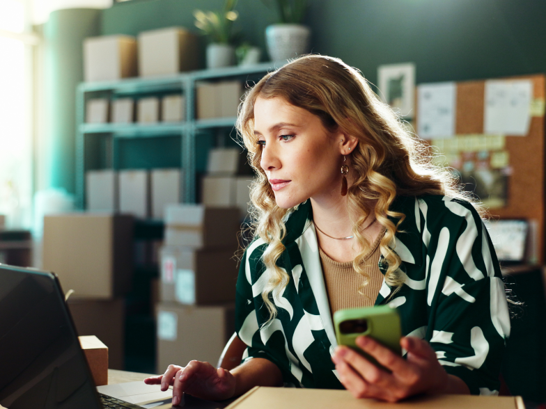 Small business owner checking her phone and laptop in a home office stocked with shipping boxes