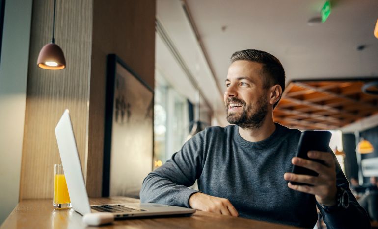 Bearded man smiling and holding a smartphone in front of a laptop