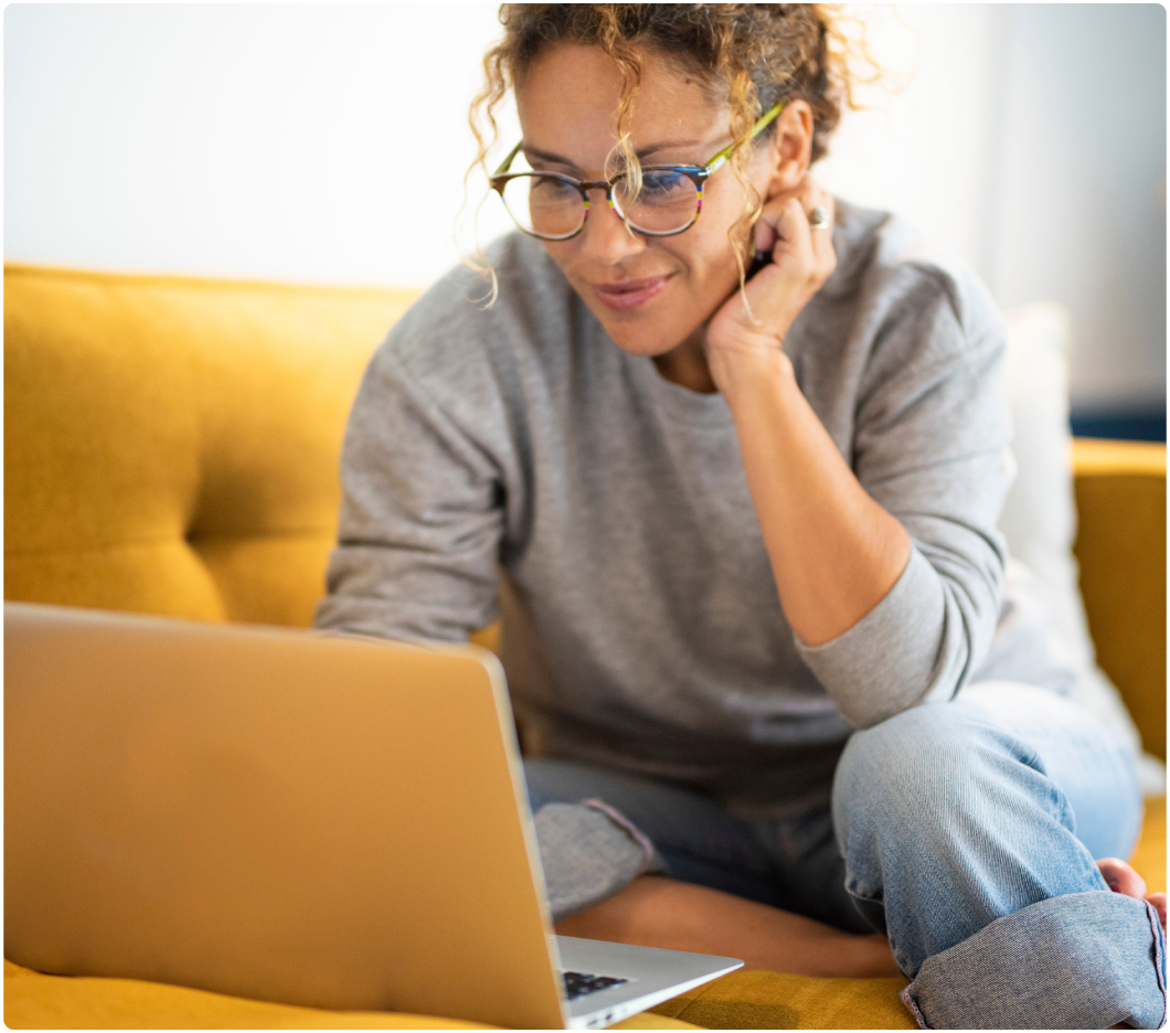 A person with curly hair and glasses, wearing a gray sweater, sits on a yellow couch, engaged with a laptop. The mood is focused and relaxed.