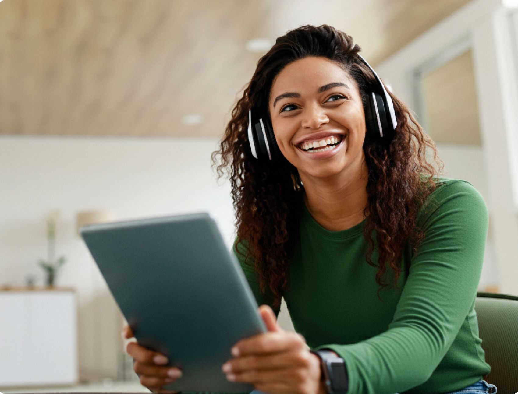 Smiling woman wearing headphones and a green shirt looks to the side while holding a laptop. She is seated in a bright room, creating a joyful atmosphere.