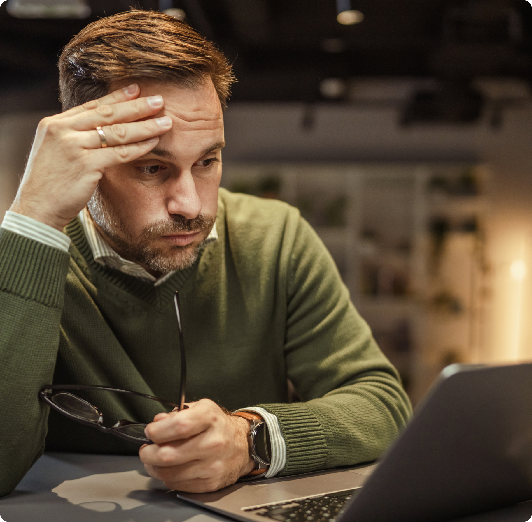 A man in a green sweater looks stressed while holding his glasses and staring at a laptop. He rests his head on his hand in a dimly lit office.