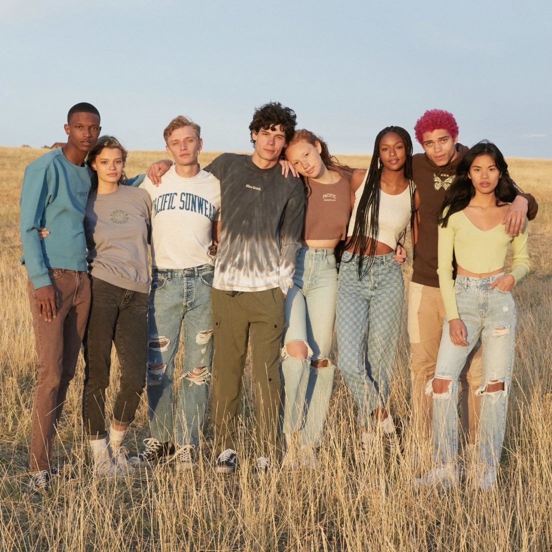 A diverse group of eight young adults stands closely together in a dry grass field, smiling under a clear blue sky, conveying a sense of friendship and unity.