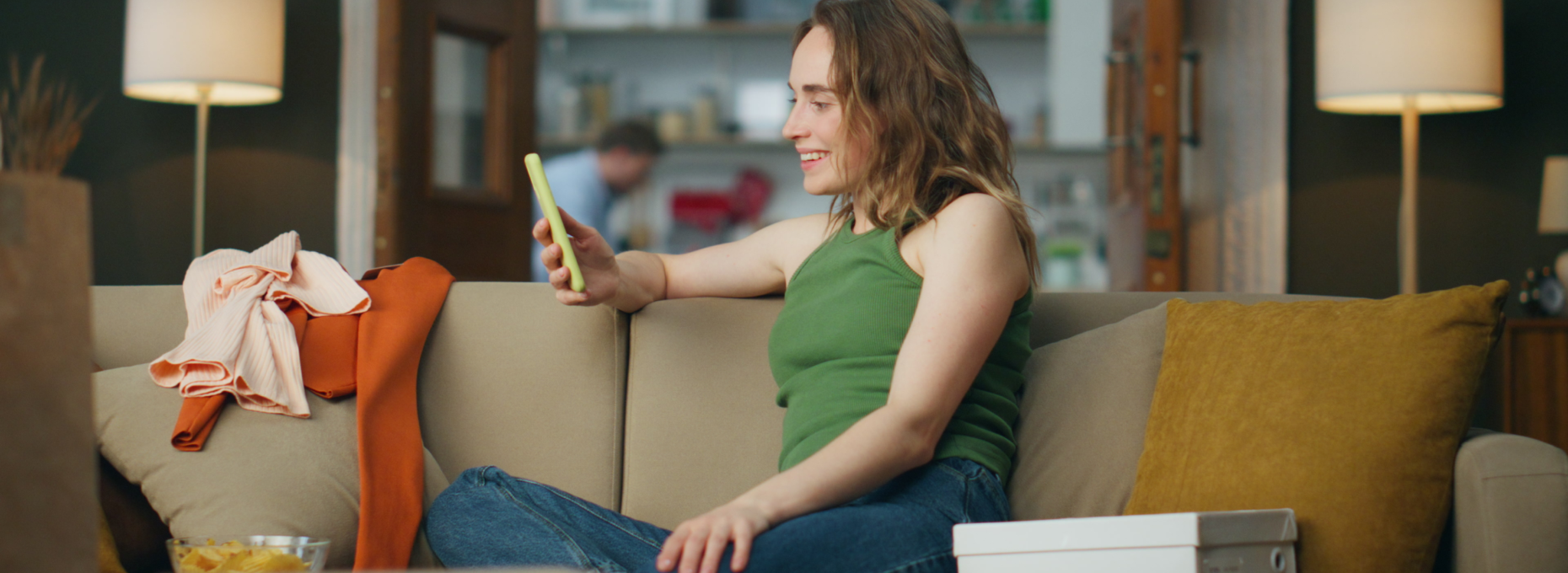A woman in a green tank top sits on a living room couch in the evening, smiling at her phone. Lamps light the room and a few clothes are draped over the arm of the couch.