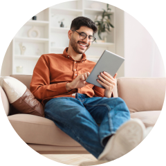 Man in glasses and orange shirt sits on beige sofa, smiling while using a tablet. Background shows white shelves with decor, creating a cozy atmosphere.