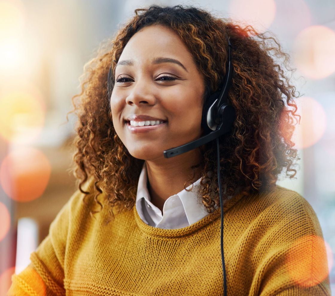 Smiling woman wearing a headset, sitting in a well-lit office. Her curly hair frames her face, and she's dressed in a mustard sweater, creating a warm atmosphere.