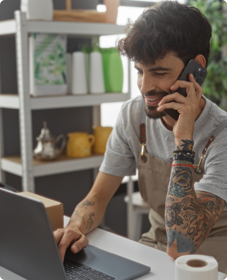 Young man with tattoos and an apron smiles while talking on the phone at a laptop. He is in a shop surrounded by colorful boxes and plants, conveying a busy yet cheerful atmosphere.