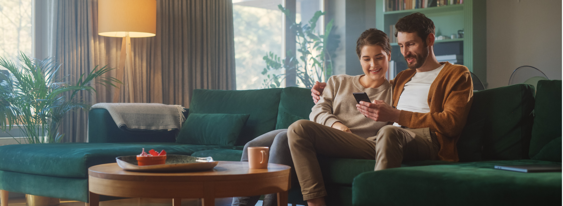 Couple relaxing on a green sofa in a sunlit living room, shopping together on a smartphone