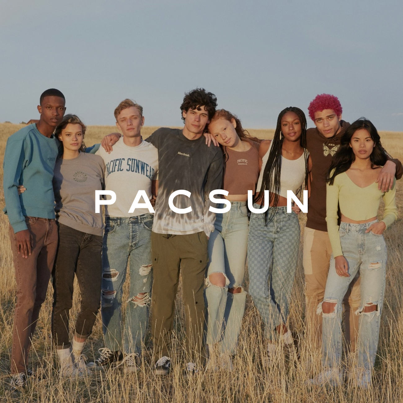 A diverse group of eight people stand closely in a sunlit field, wearing casual PacSun clothing. They appear relaxed and content, embodying a youthful vibe.