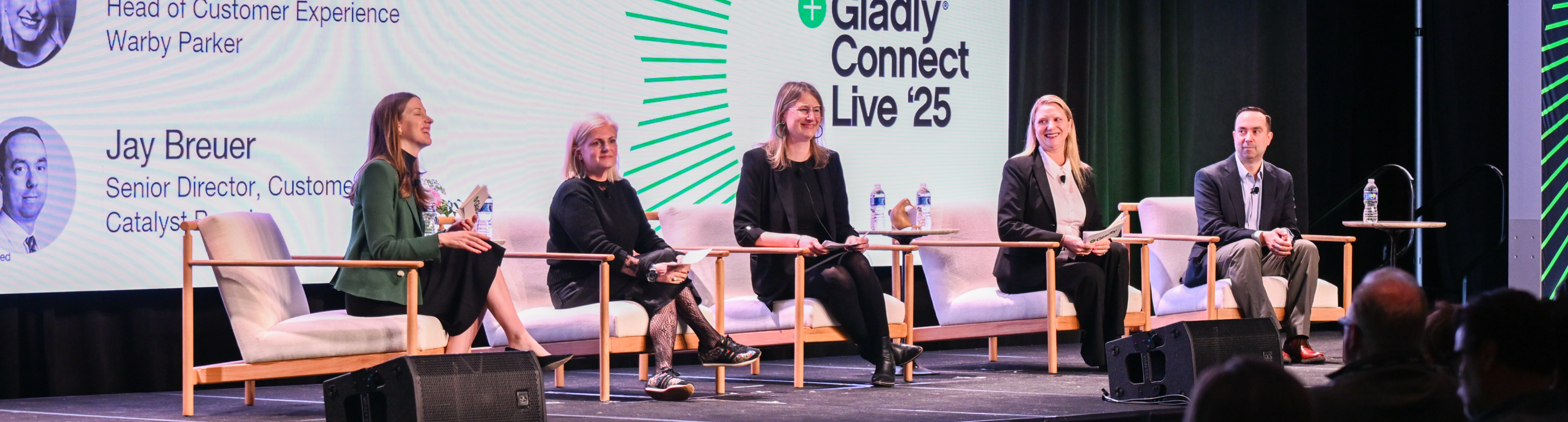 Multiple people engaged in a panel discussion on stage. One gestures while speaking; the other listens attentively, smiling. "Live '25" text is visible.