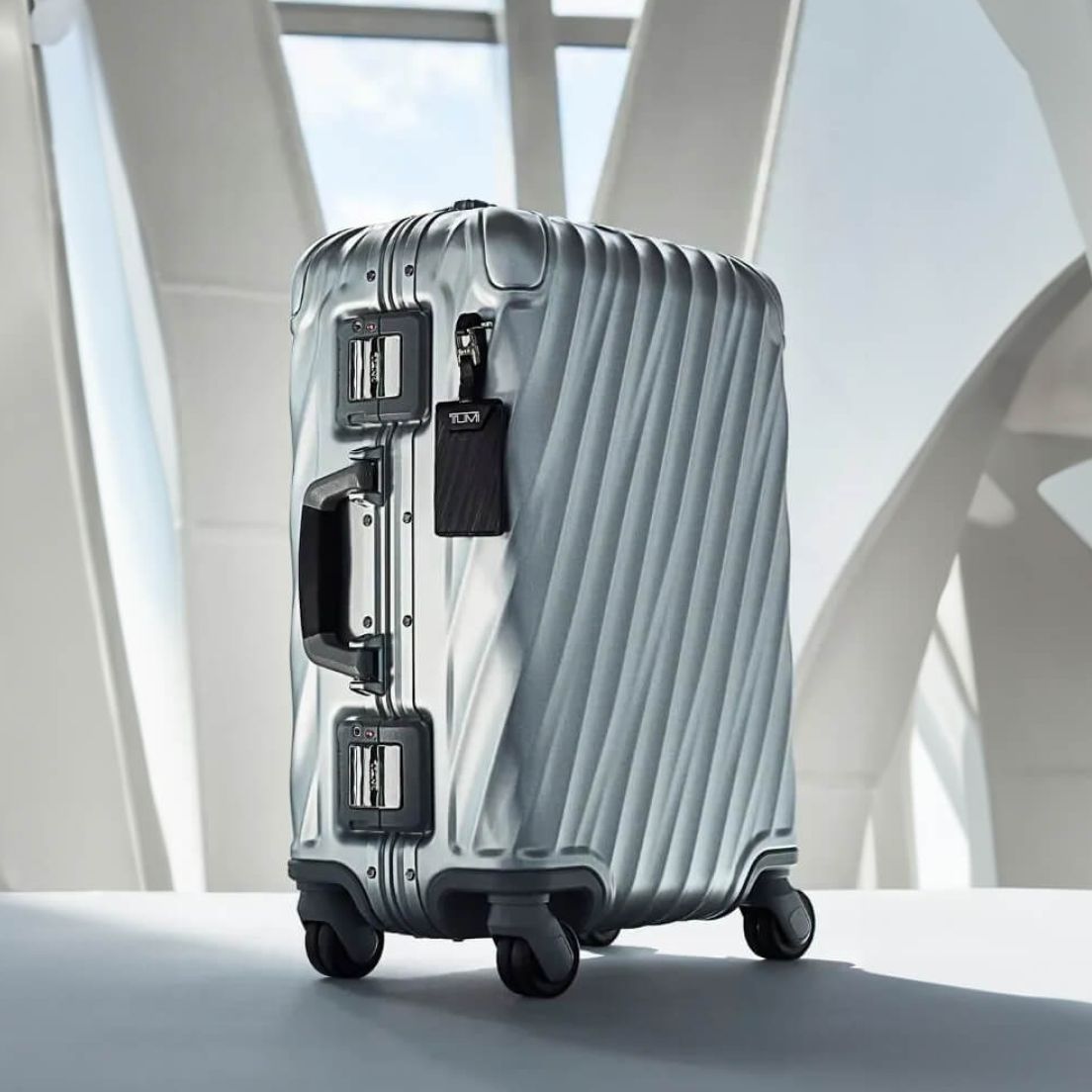 A sleek silver suitcase with a textured surface and black handles stands on four wheels. It’s set against a modern architecture backdrop, suggesting travel.