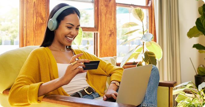 A woman in a yellow sweater and headphones smiles while online shopping with a credit card and laptop, sitting in a cozy chair by a sunlit window.