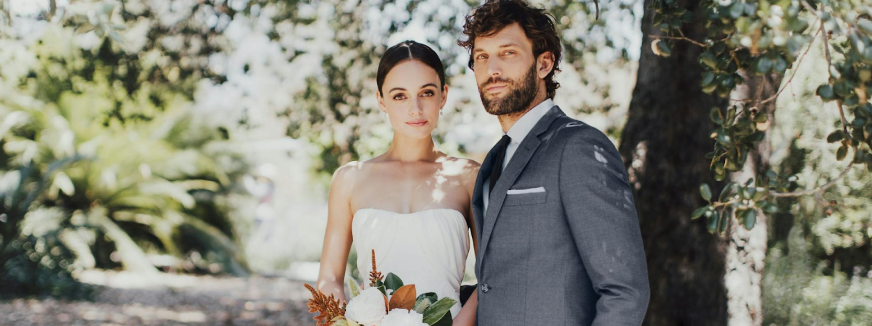 A bride in a strapless white dress holds a bouquet, standing beside a groom in a gray suit and tie. They are under a tree, exuding a serene and elegant vibe.