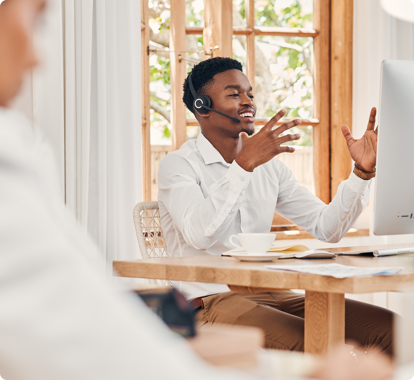 Young professional wearing a headset, smiling and gesturing while on a video call at a desk with a computer and coffee. Bright, airy office setting.