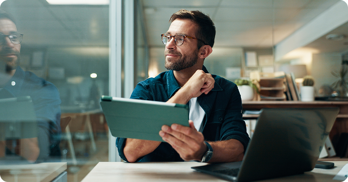 Smiling man with glasses using a laptop in a bright, plant-filled office. The scene conveys a positive and productive work atmosphere.