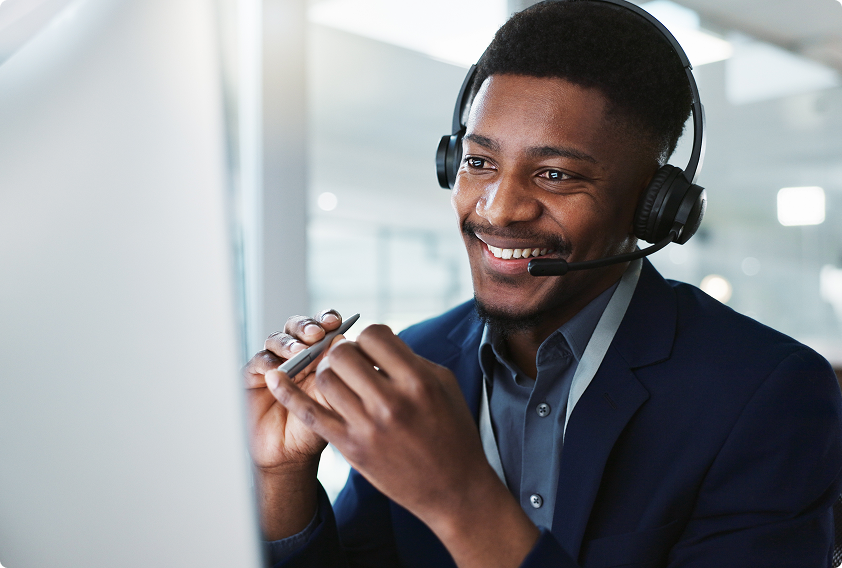 Smiling man wearing a headset and holding a pen, engaged in a video call at a desk. The setting is modern and bright, conveying a professional tone.