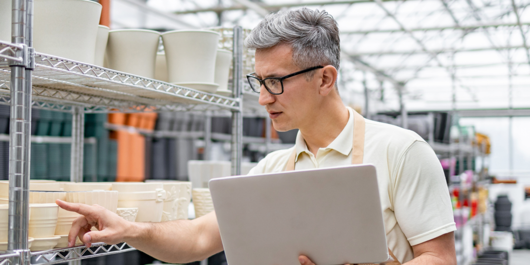 An older man in an apron and glasses holds a tablet while examining a shelf of potted plants in a greenhouse, conveying focus and professionalism.